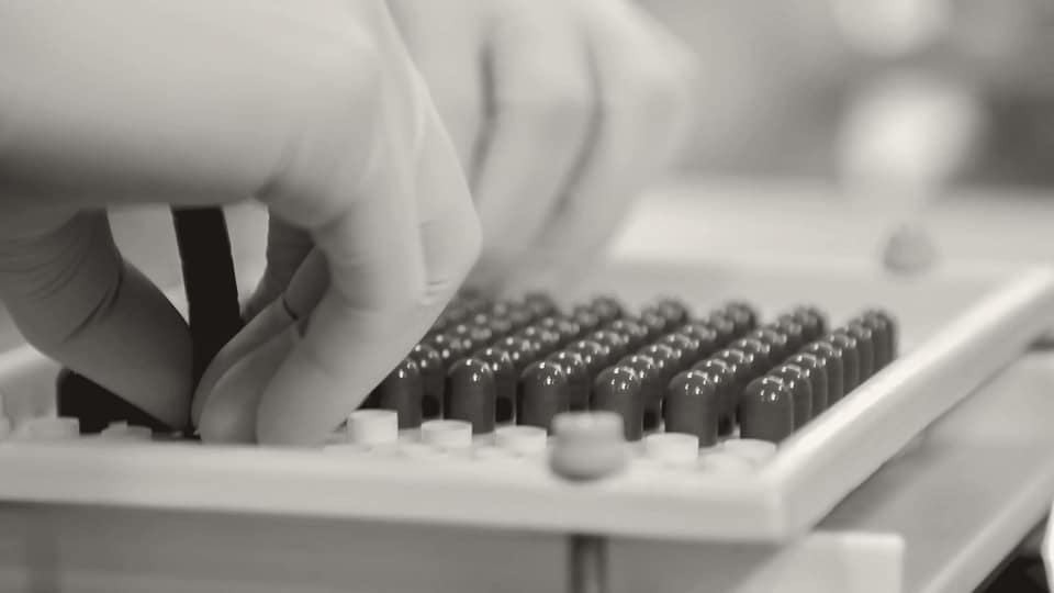 Gloved hands working with capsules in a pharmaceutical laboratory setting.